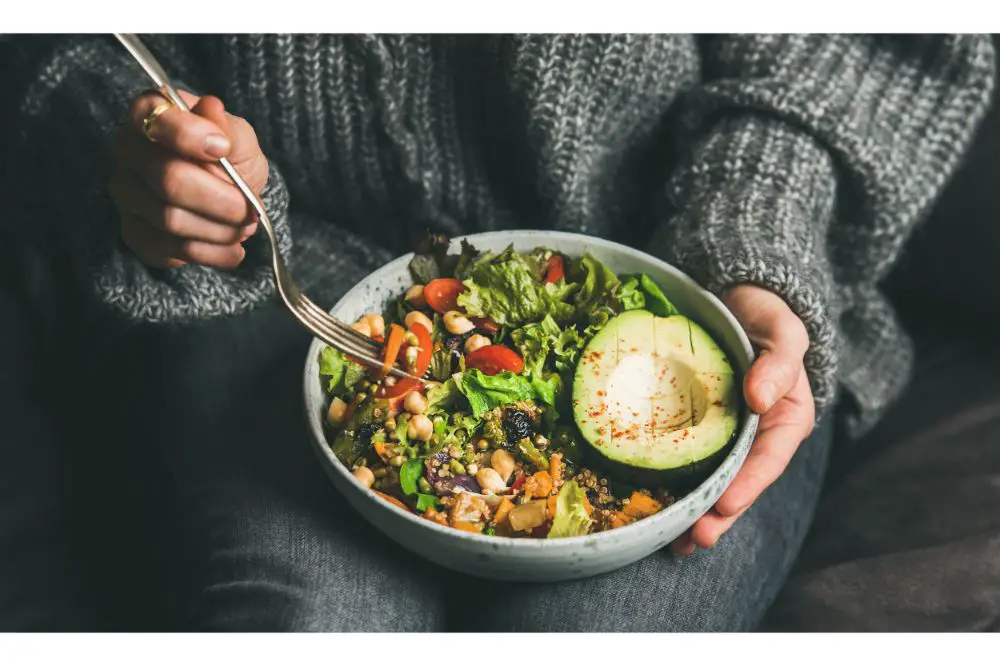 Woman in jeans and warm sweater holding bowl with fresh salad, avocado, grains, beans, roasted vegetables