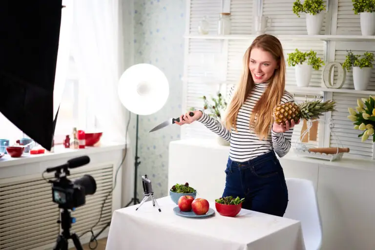 Food blogger cooking fresh vegan salad of fruits in kitchen studio, filming tutorial on camera for video channel. Female influencer holds apple, pineapple and talks about healthy eating