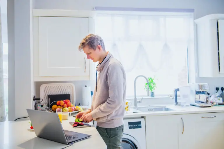 Young caucasian man working from home. Freelancer cooking vegetarian lunch on the kitchen table and working on pc laptop. Home office, remote workplace concept. Soft selective focus. Copy space